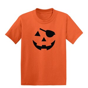May include: Orange t-shirt featuring a black jack-o'-lantern face with an eye patch. The design includes triangular eyes and a wide, smiling mouth. The shirt is a solid orange color.