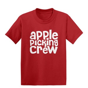 以下が含まれることがあります： 赤い半袖Tシャツに、白い文字で「apple picking crew」と書いてあり、小さなリンゴのグラフィックがあります。