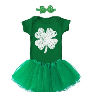 May include: A green baby outfit with a white four-leaf clover graphic. The outfit includes a green tutu and a green bow headband.