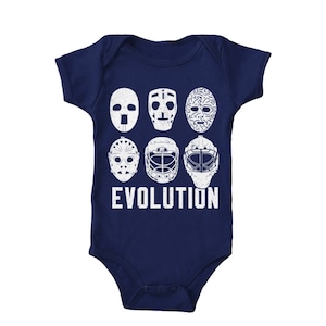May include: A navy blue baby onesie with a white graphic of hockey goalie masks, showing the evolution of the mask design. The text "EVOLUTION" is printed below the masks.