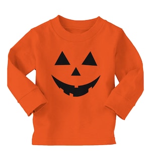 Puede incluir: Camiseta naranja de manga larga con una cara de calabaza negra. El diseño incluye ojos triangulares, una nariz triangular y una boca sonriente. La camiseta está hecha de un material suave, perfecta para Halloween.