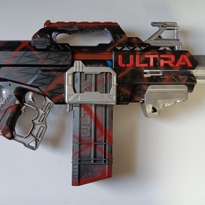 May include: A black and red toy blaster with the word "ULTRA" in red lettering. The blaster has a handle, a magazine, and various silver-colored accents. The word "NERF" is visible on the magazine.