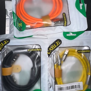 Puede incluir: Tres paquetes de cables de carga de diferentes colores. El primer paquete contiene un cable naranja, el segundo un cable negro y el tercero un cable amarillo. Cada paquete tiene la palabra "Cable" impresa, junto con las palabras "Alta calidad" y "Accesorios".