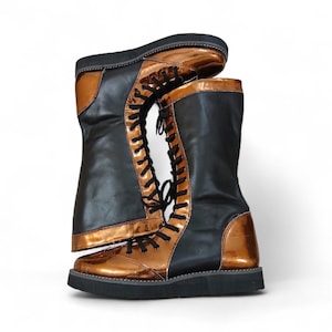 Puede incluir: Un par de botas negras y bronce con un acabado brillante. Las botas tienen un diseño con cordones con cordones negros y una suela gruesa negra. Las secciones de bronce están en la puntera, el talón y los bordes superiores.