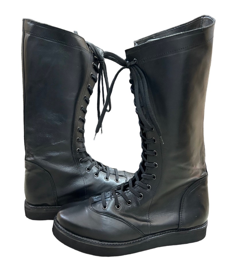 Puede incluir: Un par de botas de lucha libre de cuero negro con cordones. Las botas tienen una suela gruesa y son de altura de rodilla.