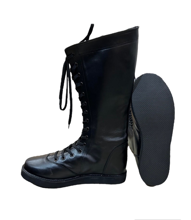Puede incluir: Botas de lucha libre de cuero negro con cordones en el lateral y suela gruesa. Las botas se muestran con la suela de una bota mirando hacia la c&aacute;mara.