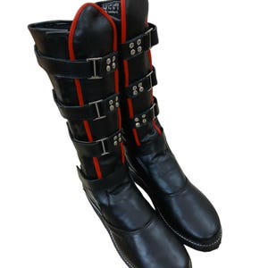 Könnte beinhalten: Schwarze Lederstiefel mit mehreren Riemen und silbernen Schnallen. Die Stiefel haben rote Akzente an den Seiten und oben sowie weiße Nähte um die Sohlen. Die Stiefel sind kniehoch.