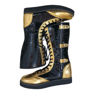 May include: Black and gold knee-high boots with a glossy finish. The boots have gold accents on the toe, heel, and upper shaft, with a lace-up front and a zip closure. The soles are black.