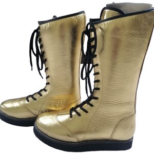 May include: A pair of gold wrestling boots with black laces. The boots are made of leather and have a thick sole.