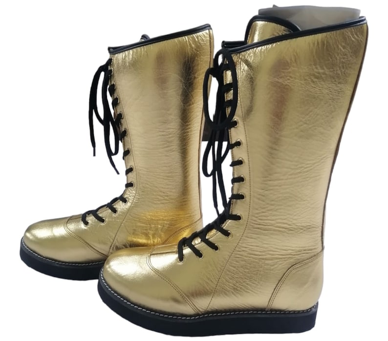 May include: A pair of gold wrestling boots with black laces. The boots are made of leather and have a thick sole.