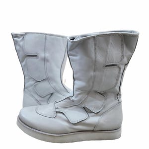 Könnte beinhalten: Ein Paar weiße Lederstiefel mit einem einzigartigen Design mit Ausschnitten und Riemen. Die Stiefel haben eine dicke Sohle und einen hohen Schaft.
