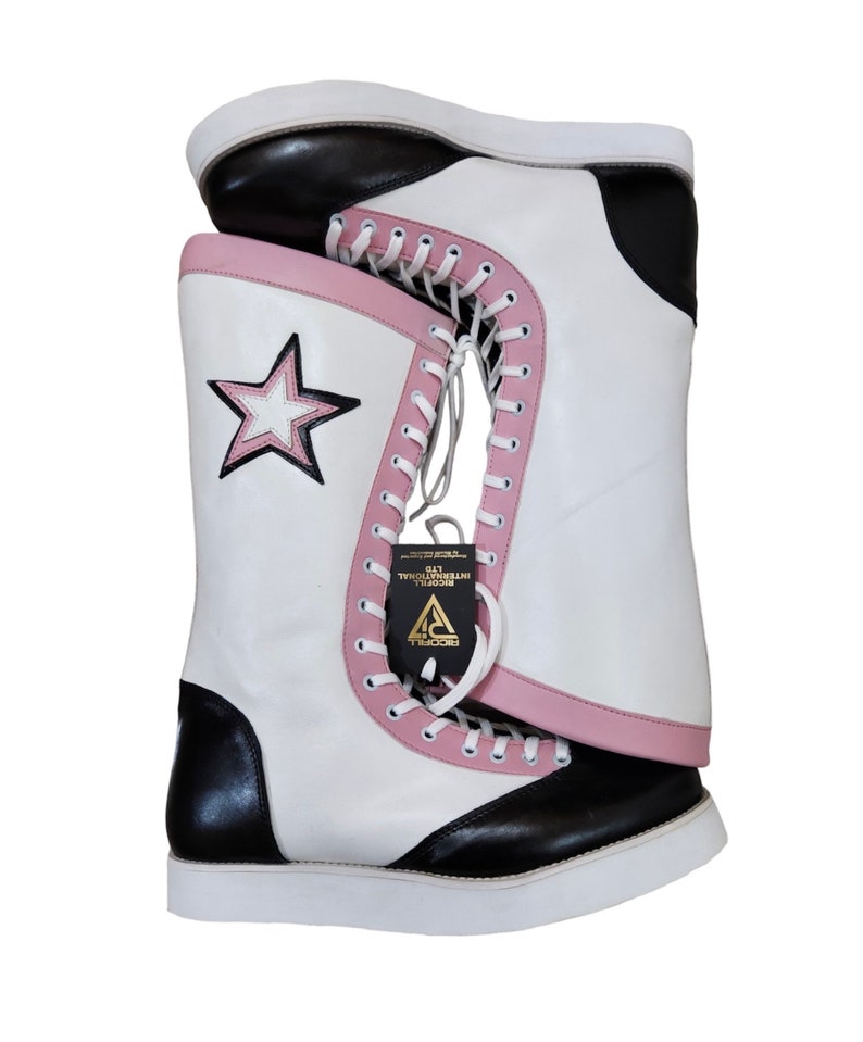 May include: White leather wrestling boots with black accents and pink trim. The boots have a star design on the side and a white lace-up closure. The boots are sitting on a white surface.