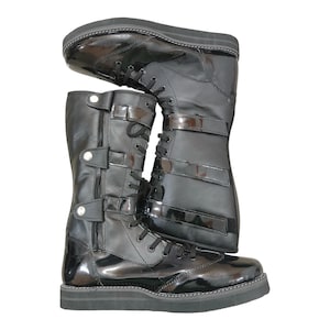 Könnte beinhalten: Paar schwarze, kniehohe Stiefel mit glänzendem Finish. Die Stiefel haben mehrere Riemen mit Schnallen und Schnürsenkeln sowie eine dicke, schwarze Sohle. Das Design erinnert an Kampfstiefel, mit einer modernen, eleganten Ästhetik.