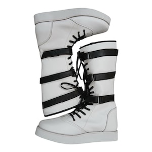 Könnte beinhalten: Weiße Stiefel mit schwarzen Riemen und Schnürsenkeln. Die Stiefel haben eine flache Sohle und eine mittlere Wadenhöhe. Das Design zeichnet sich durch eine Kombination aus glattem weißem Material und schwarzen Akzenten aus.