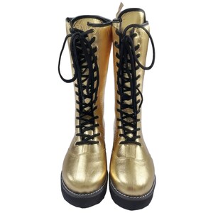 May include: A pair of gold leather wrestling boots with black laces. The boots have a thick sole and are laced up the front.
