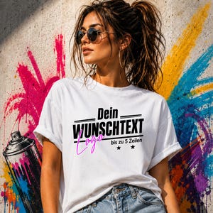 Könnte beinhalten: Ein weißes T-Shirt mit dem schwarzen Text "Dein WUNSCHTEXT", dem Wort "Logo" in pinker Schreibschrift darunter, gefolgt von "bis zu 5 Zeilen" und zwei kleinen schwarzen Sternen. Das T-Shirt wird zu einer blauen Jeans getragen.