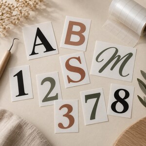 Könnte beinhalten: Eine Sammlung einzelner selbstklebender Vinyl-Aufkleber mit verschiedenen Buchstaben und Zahlen in Schwarz, Olivgrün und Terrakotta. Das Set enthält die Zeichen A, B, S, M, 1, 2, 3, 7 und 8 auf weißem, rechteckigem Trägerpapier gedruckt.