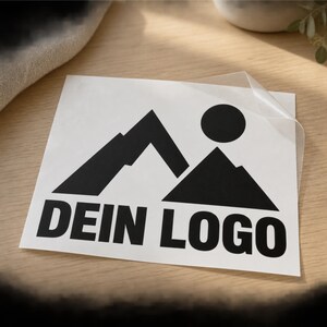 Könnte beinhalten: Ein quadratischer weißer Aufkleber mit einer schwarzen Grafik aus zwei Berggipfeln und einer runden Sonne. Unter der Grafik ist der Text DEIN LOGO in einer fetten, schwarzen, serifenlosen Schrift gedruckt. Der Aufkleber hat eine transparente Transferfolie.
