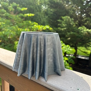 May include: A gray, textured planter with a unique, draped design. The planter has a wide opening and a flared, pleated skirt-like shape. The color is a muted gray, and the planter is sitting on a wooden deck.