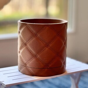 May include: A cylindrical brown container with a quilted diamond pattern. The container is made of a leather-like material and sits on a white table. The background is blurred, suggesting an indoor setting.