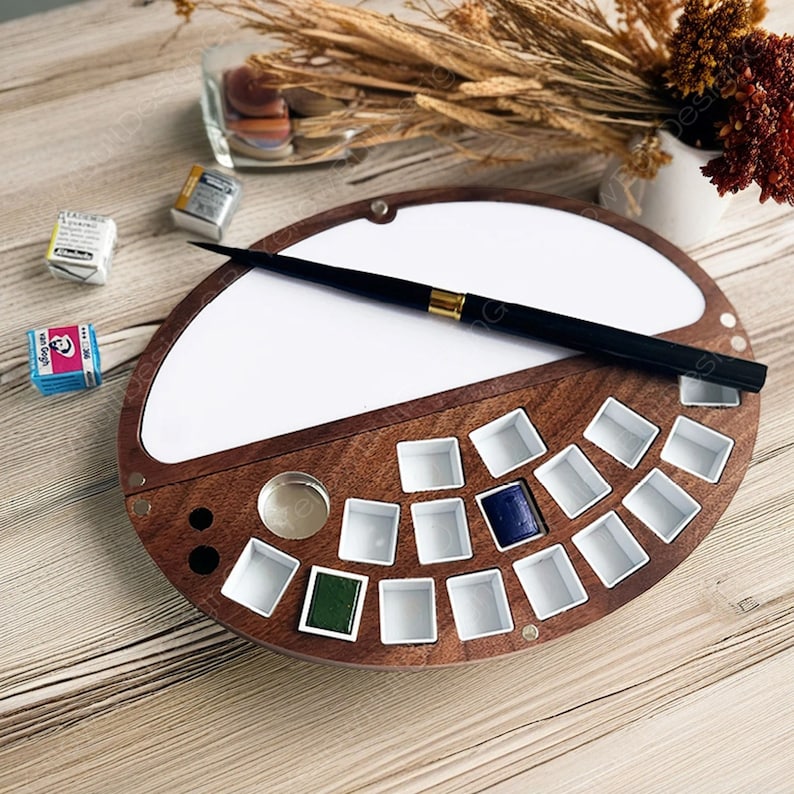Op de afbeelding: Een houten aquarelpalette met een wit deksel en 18 vakjes voor verf. De palette heeft een ingebouwde penseelhouder en een klein metalen bakje voor water. Een zwart penseel ligt op het deksel.