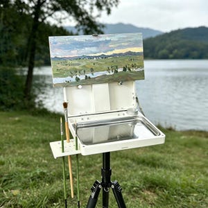 May include: A white portable plein air easel with a landscape painting. The easel has a metal tray, brush holders, and is mounted on a black tripod. The painting depicts a river, trees, and a cloudy sky.