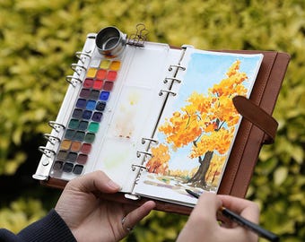May include: A brown leather-bound watercolor art journal with a built-in palette and a watercolor painting of a tree with yellow and orange leaves. The journal is open, revealing the painting and the palette with various colors.