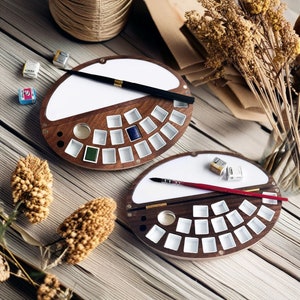 Op de afbeelding: Twee ronde houten aquarelpaletten met deksels die opengaan om rijen lege verfputjes te onthullen. Elke palet heeft een wit oppervlak om verf te mengen. Een palet heeft een penseel dat op het witte oppervlak ligt.