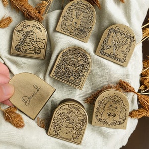May include: A selection of antique brass-coloured metal book corners, each with an engraved design. Designs include a rabbit, a cat, a butterfly, a unicorn, and the words "Love Letter" and "Obscure Love". One corner is engraved with the name "David".