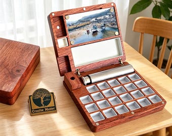 May include: A wooden watercolor paint box with a hinged lid, open to reveal a palette of small, square paint wells. The box is made of reddish-brown wood and includes a metal tube and a small picture of a coastal scene.