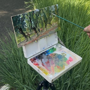 Könnte beinhalten: Ein Künstler-Pleinair-Setup. Ein kleines Gemälde einer Parkszene ist auf einer weißen Staffelei montiert, darunter eine Palette mit gemischten Farben. Ein Pinsel wird über dem Gemälde gehalten. Das Setup steht auf einem Stativ, umgeben von grünem Gras.