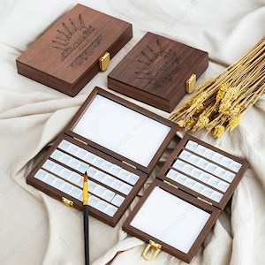 Op de afbeelding: Een houten aquarelverfdoos met een scharnierend deksel. Het deksel is gegraveerd met een zon, maan en kwasten. De doos is open en toont de lege verfpotjes. De doos staat op een wit stoffen oppervlak.