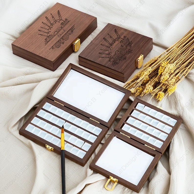 Op de afbeelding: Een houten aquarelverfdoos met een scharnierend deksel. Het deksel is gegraveerd met een zon, maan en kwasten. De doos is open en toont de lege verfpotjes. De doos staat op een wit stoffen oppervlak.