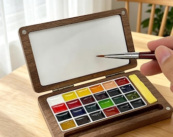 May include: A wooden watercolor paint set with a hinged lid, featuring a white mixing surface and a palette of vibrant colors. A paintbrush is poised above the palette, ready for use. The set is open on a wooden table.