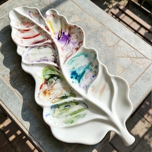 Könnte beinhalten: Weißes Keramik-Serviertablett in Blattform mit farbenfrohen abstrakten Aquarelldesigns. Das Tablett hat mehrere Fächer und eignet sich ideal zum Servieren von Vorspeisen oder zum Präsentieren von Dekorationsartikeln. Die Designs umfassen Rot-, Lila-, Grün- und Blautöne.