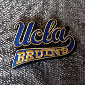 May include: An embroidered patch featuring the text Ucla in blue script with a gold border, positioned above the word BRUINS in gold block letters on a blue background with a black border, creating a layered collegiate sports emblem design.