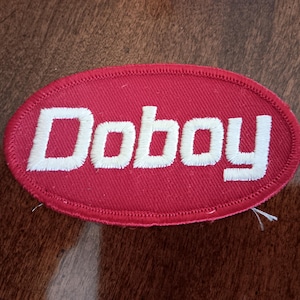 May include: Red oval patch with the word "Doboy" embroidered in white. The patch has a stitched border and is set against a brown and white patterned background. The patch is likely for clothing or accessories.
