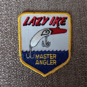 May include: Embroidered patch with a white, red, and black fishing lure design. The patch has the words "LAZY IKE" in red and "MASTER ANGLER" in yellow. The patch is bordered in yellow and blue.
