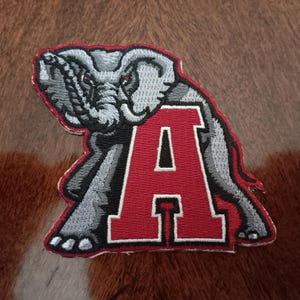 May include: Embroidered patch featuring a gray elephant with red eyes and a red letter "A". The elephant is positioned over the letter, which has a white outline. The patch is on a brown and tan textured surface.