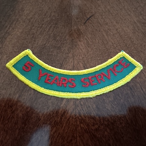 May include: A green and yellow embroidered patch with the words "5 YEARS SERVICE" in red. The patch has a curved shape and is likely for a uniform or garment.