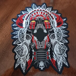 May include: An embroidered patch featuring a skull wearing a feathered headdress in red, white, gray, and black. The skull has glowing yellow eyes and an open mouth, with a smaller skull icon at the bottom and intricate feather detailing throughout.