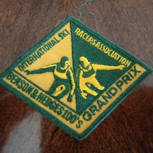 May include: A diamond-shaped embroidered patch featuring a split yellow and green background. It depicts two skiers in yellow and includes the text INTERNATIONAL SKI RACERS ASSOCIATION and BENSON & HEDGES 100'S GRAND PRIX in yellow and green.