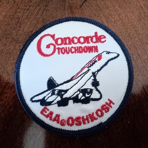 May include: A circular embroidered patch with a white background and a dark blue border. It features a black and white illustration of a Concorde airplane, with the red text "Concorde TOUCHDOWN" above and "EAA OSHKOSH" below the aircraft.