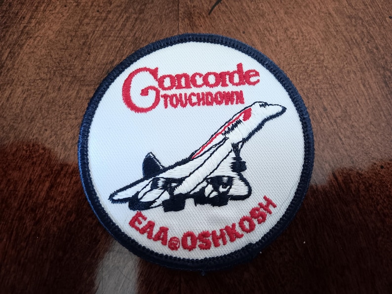 May include: A circular embroidered patch with a white background and a dark blue border. It features a black and white illustration of a Concorde airplane, with the red text "Concorde TOUCHDOWN" above and "EAA OSHKOSH" below the aircraft.