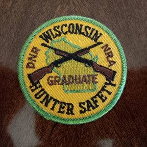 May include: A circular embroidered patch with a yellow background and a green border. It features a green outline of the state of Wisconsin, two crossed rifles, and black text reading DNR WISCONSIN NRA GRADUATE HUNTER SAFETY.