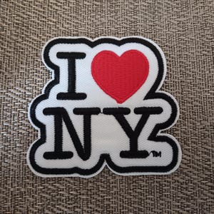 May include: Embroidered patch featuring the phrase "I NY" with a red heart replacing the word "love". The patch has a white background with black lettering and a black border. The patch is a souvenir or a decorative item.