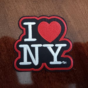 May include: An embroidered patch featuring the text I, a red heart symbol, and the letters NY in white, set against a black background with a red border. The design includes a small trademark symbol next to the letter Y, with white stitching.
