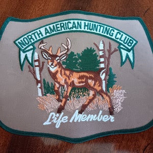 May include: A tan embroidered patch with a green border. The patch features a white banner with the text "NORTH AMERICAN HUNTING CLUB" and a white banner with the text "Life Member". The patch also features a brown and white deer standing in a green forest.