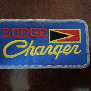 May include: Embroidered patch with the text "DODGE Charger" in red and yellow on a blue background. The patch features a stylized flag design with black, red, and yellow elements, and a gray border.