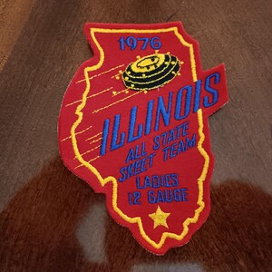 May include: A red and yellow embroidered patch in the shape of Illinois. The patch reads "ILLINOIS ALL STATE SKEET TEAM LADIES 12 GAUGE" with the year "1976" at the top. A skeet target is in the center.